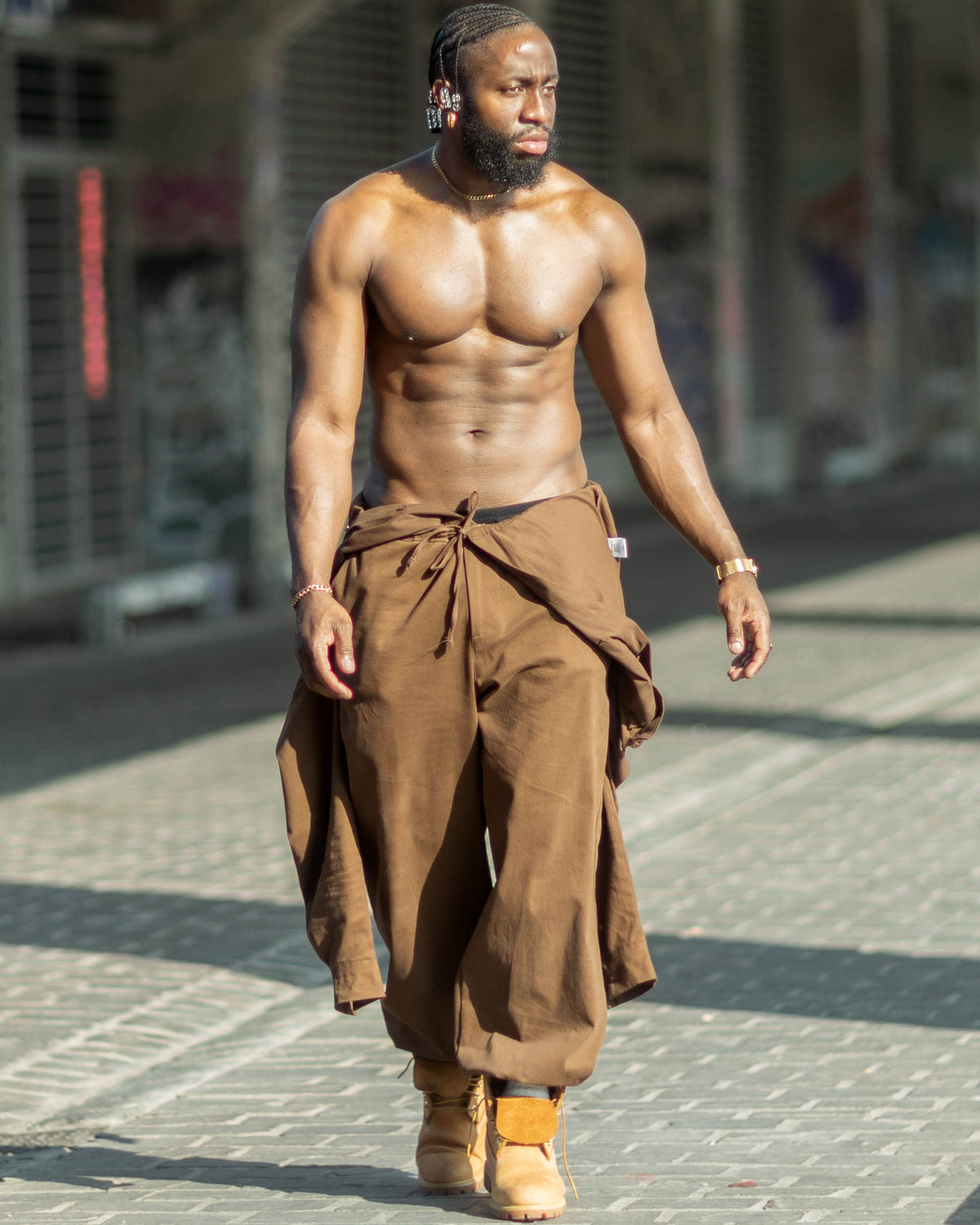 Combinaison oversize marron James Suit, haut noué autour de la taille, mannequin torse nu, bottes camel, pantalon ample resserré à la cheville